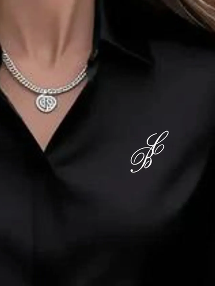 Close-up of the white monogram embroidered over the heart of the black silk shirt
